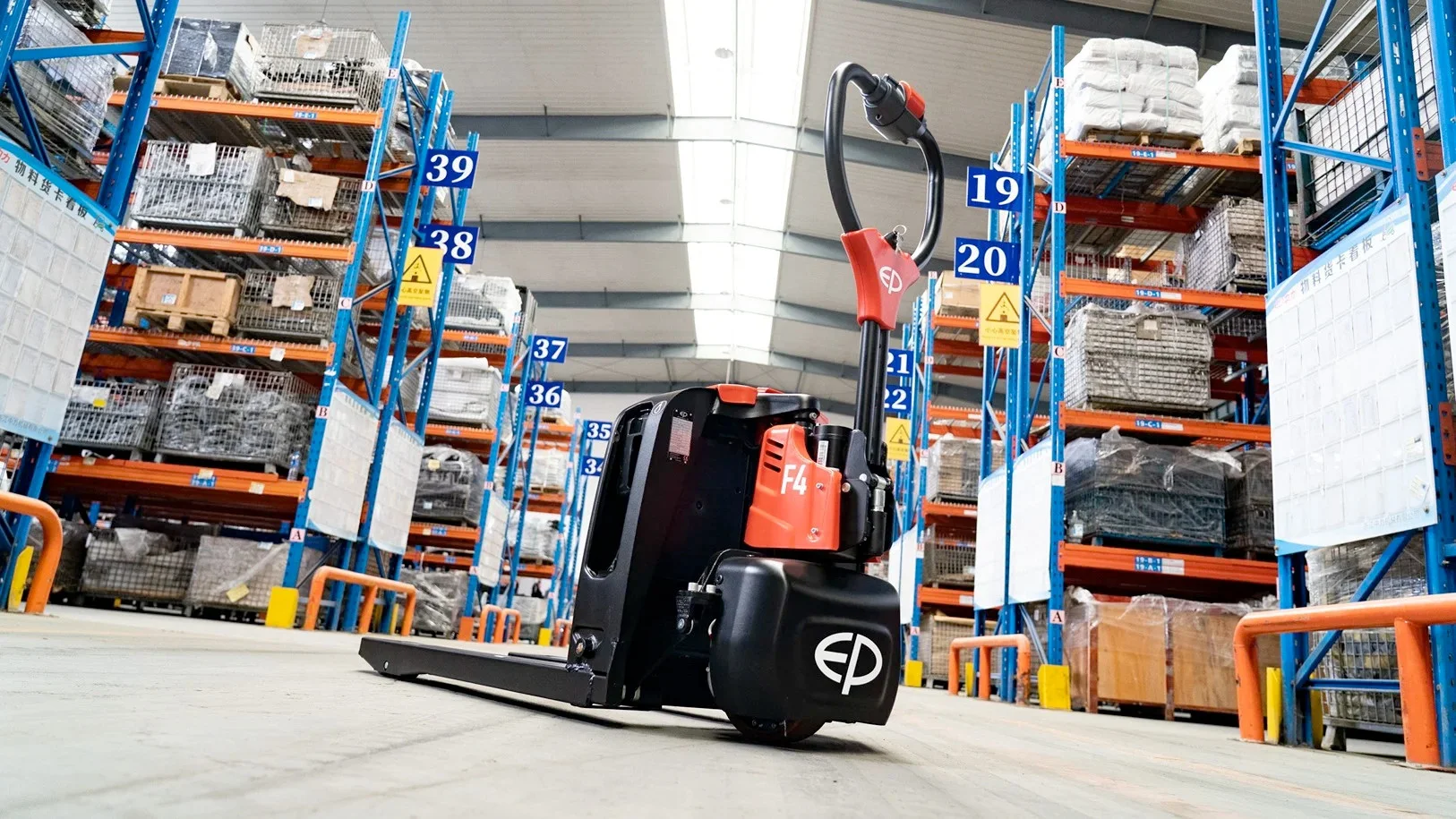 Hubwagen mittig im Vordergrund, dahinter Lagerhalle mit hohen blauen Regalen voller Kartons.