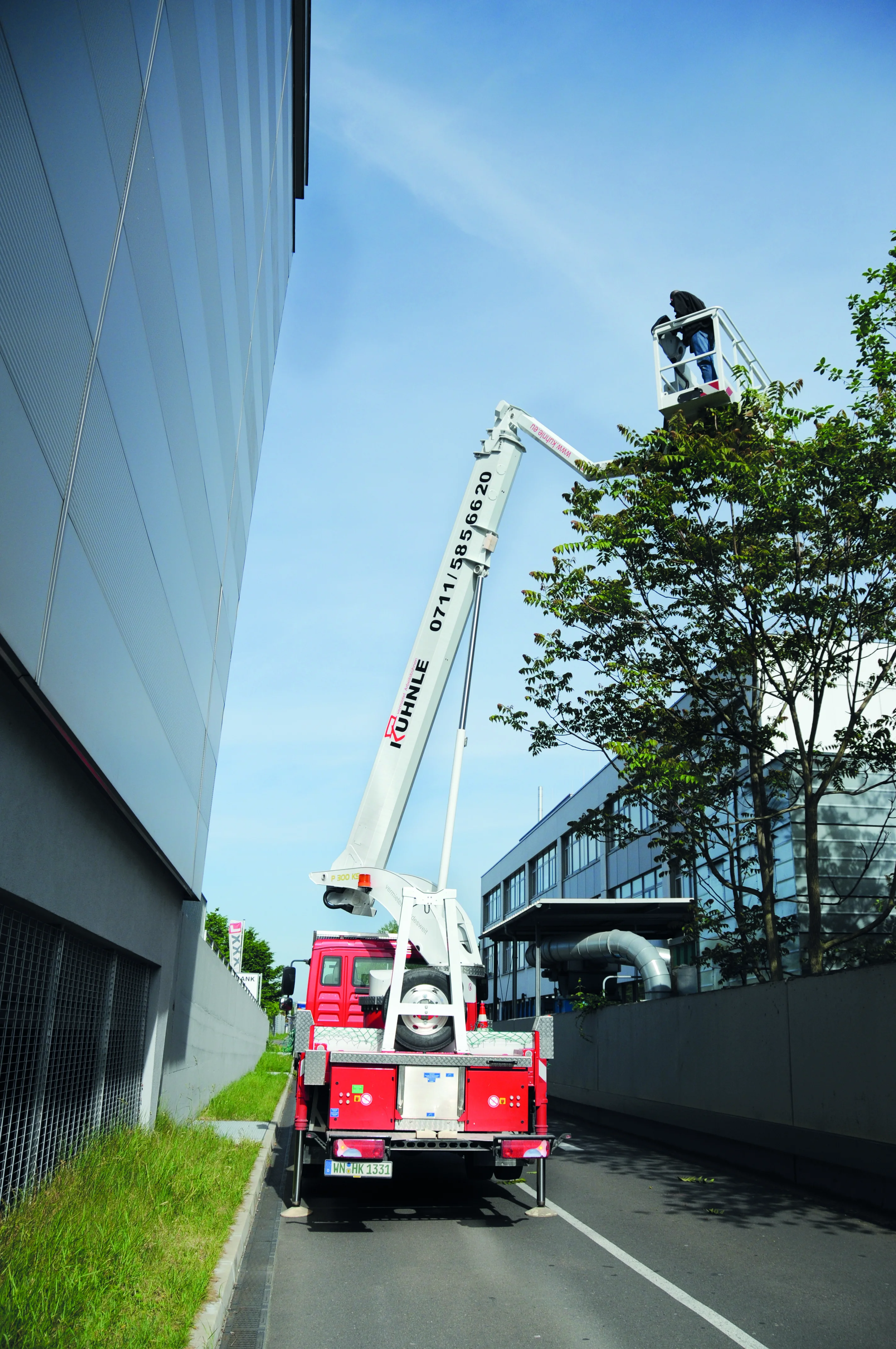 Rote ausgefahrene Arbeitsbühne zwischen zwei gläsernen Gebäuden, rechts ein Baum.