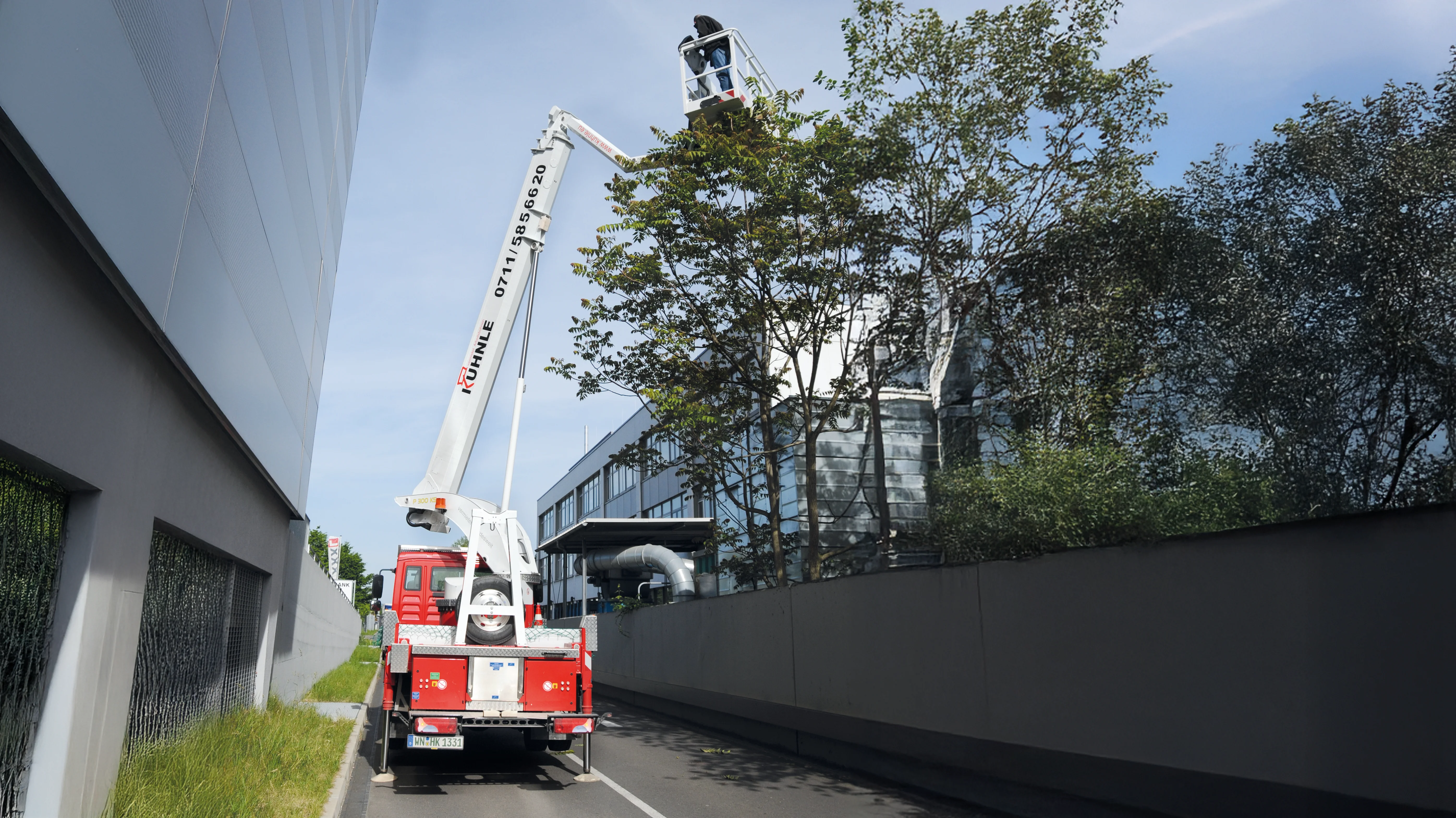 Rote ausgefahrene Arbeitsbühne zwischen zwei gläsernen Gebäuden, rechts ein Baum.