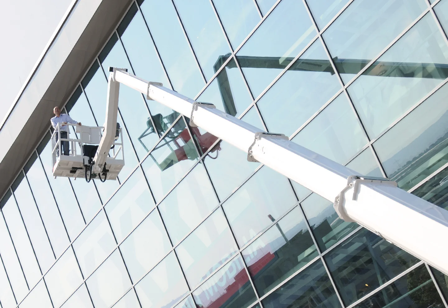 Ausgefahrene Arbeitsbühne vor hohem gläsernem Gebäude, darauf eine Person, links oben Himmel und Wolken