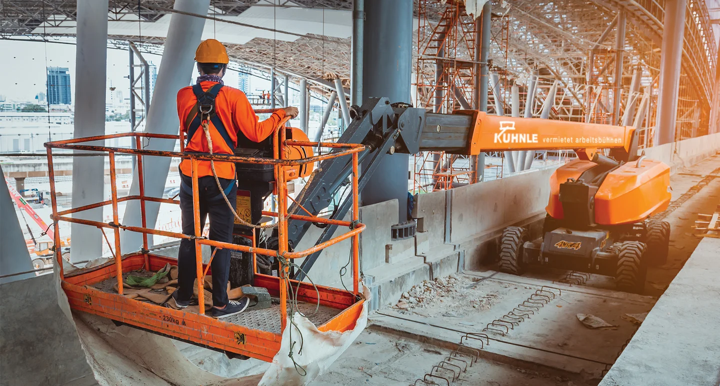 Person in Schutzkleidung auf orangener Arbeitsbühne, die sich unter überdachter Struktur mit sichtbaren Säulen befindet.