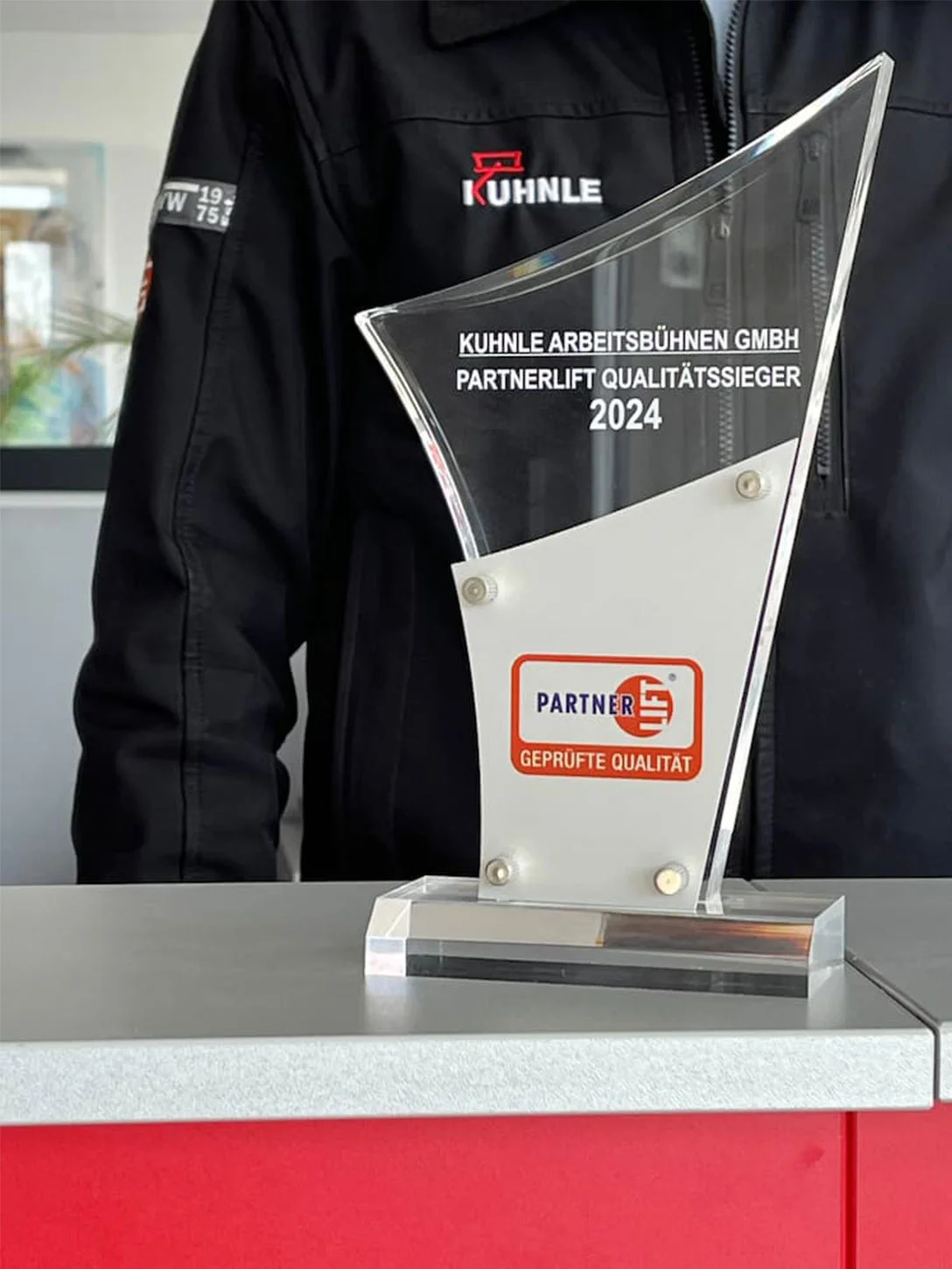 Award auf Tisch, dahinter Person in schwarzer Arbeitsjacke.