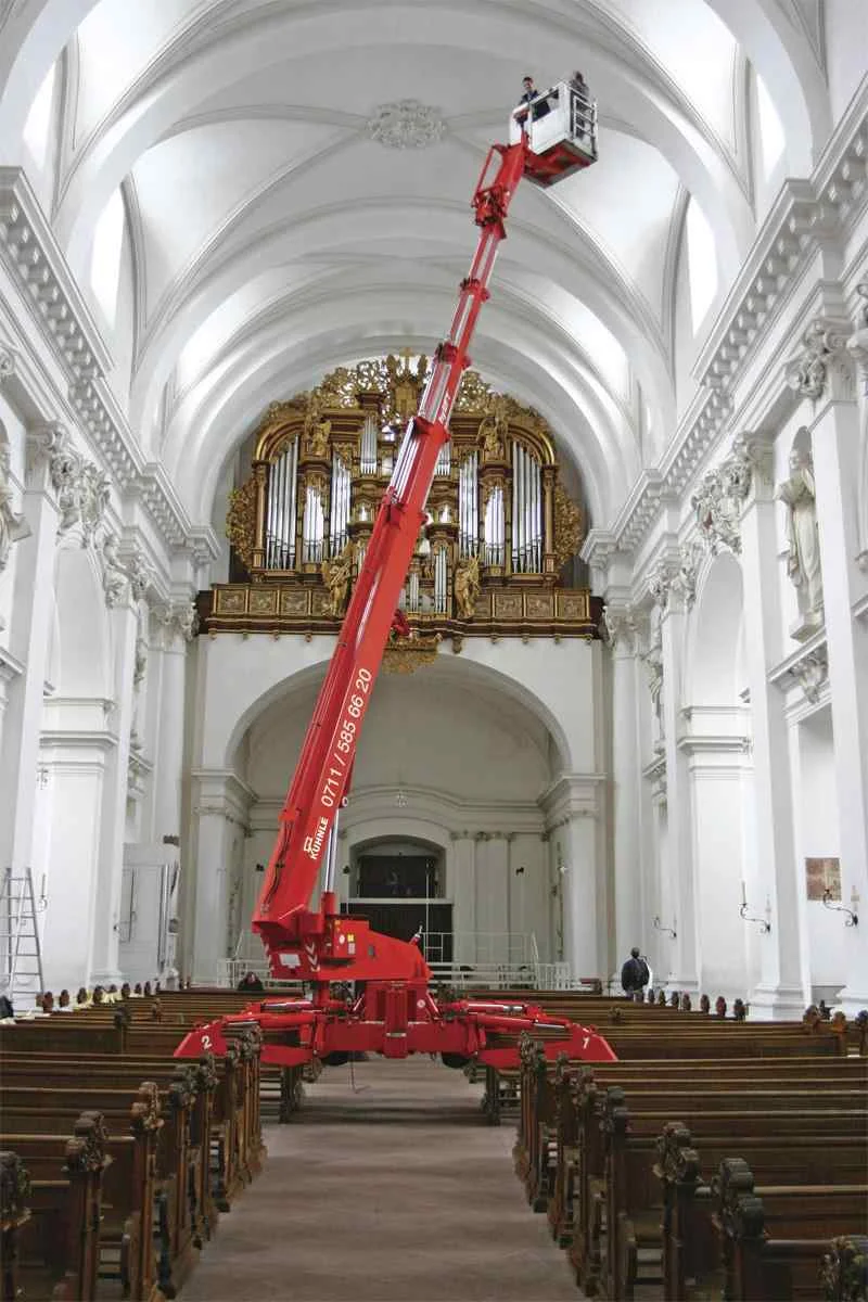 Ausgefahrene Arbeitsbühne mittig in einer Kirche, davor Sitzbänke.