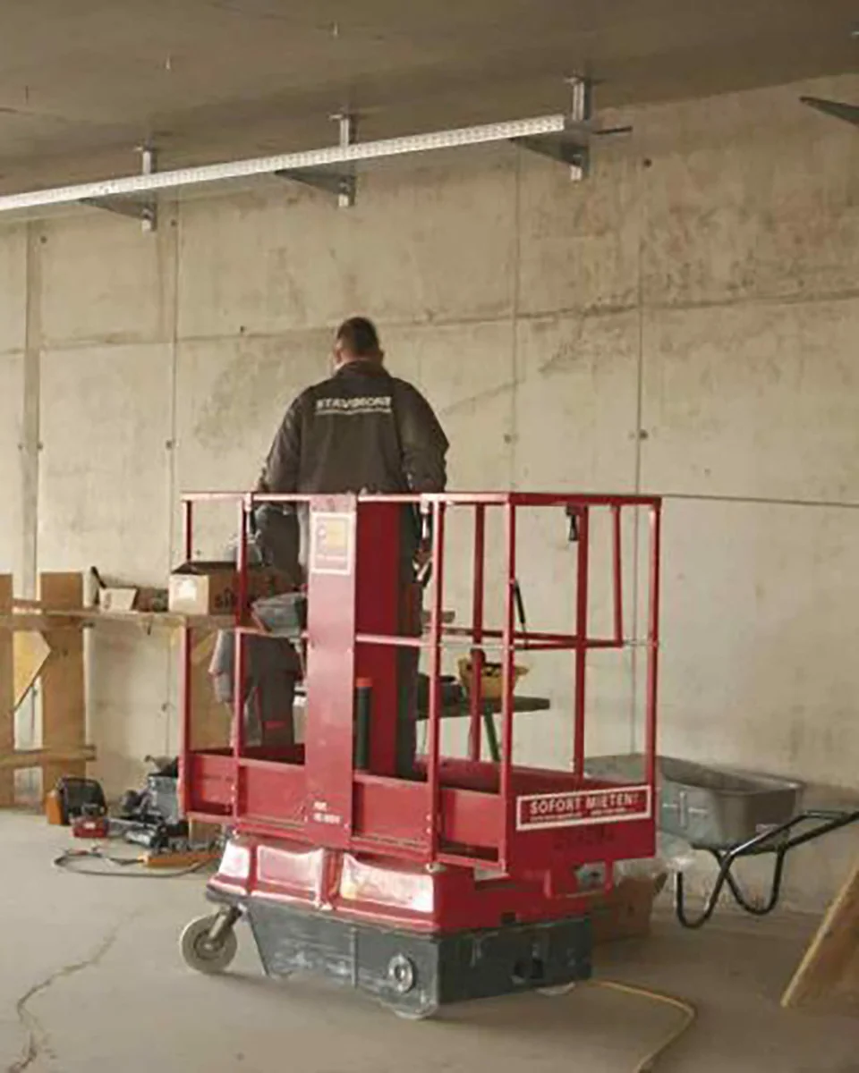 Person auf roter Arbeitsbühne auf dem Boden, in Arbeitsumgebung mit Schubkarre, hinten eine Steinwand.