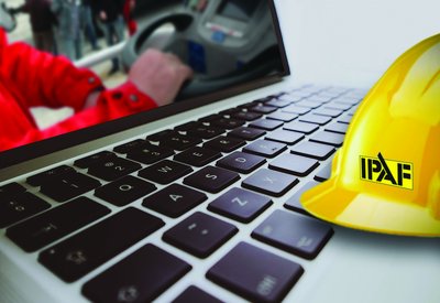 Gelber Schutzhelm liegt auf einem Laptop, dessen Bildschirm eine teilweise sichtbare Person auf einer Arbeitsbühne zeigt.