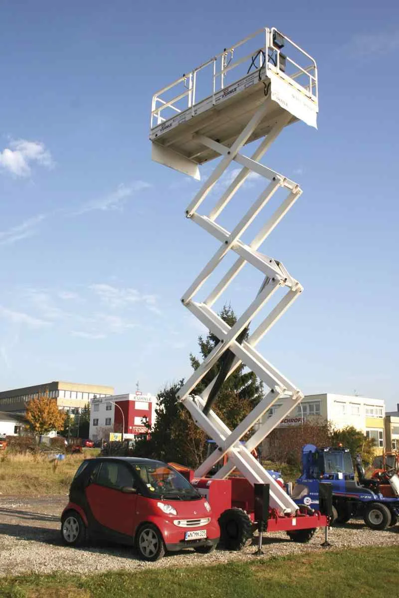 Ausgefahrene Arbeitsbühne, daneben geparkt ein roter Smart und ein blaues Arbeitsgerät, im Hintergrund weitere Fahrzeuge, Feld und Gebäude.