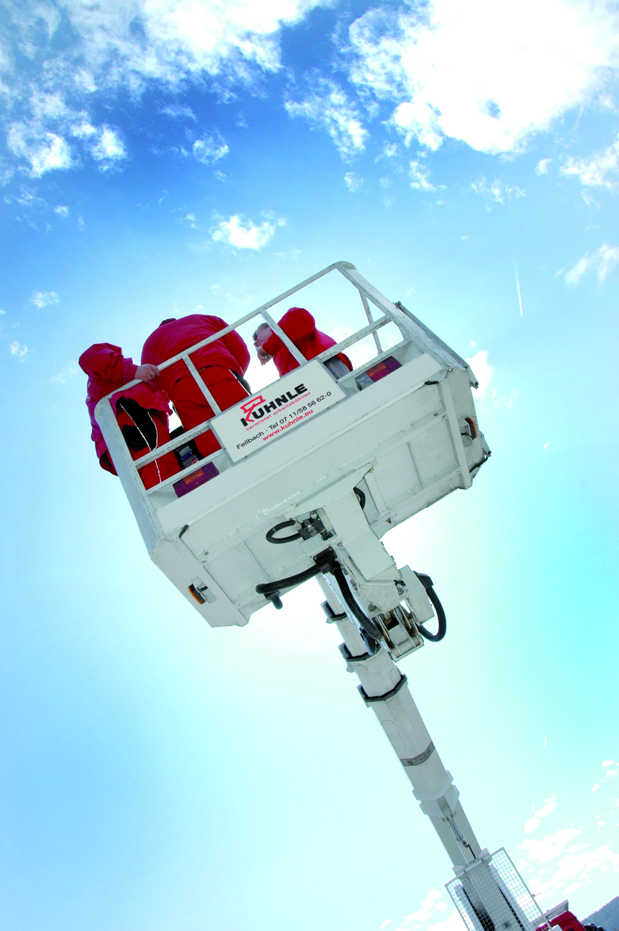 Arbeitsbühne in ausgefahrenem Zustand, darauf drei Personen in Rot, vor leicht wolkigem blauen Himmel.