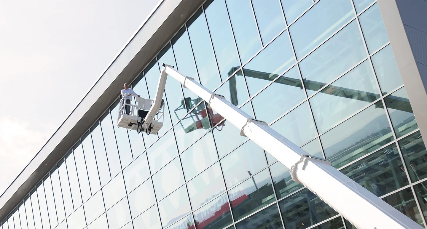 Ausgefahrene Arbeitsbühne vor hohem gläsernem Gebäude, darauf eine Person, links oben Himmel und Wolken