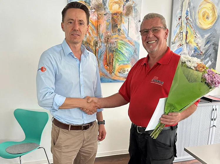 Zwei Männer geben sich die Hand, einer hält einen Blumenstrauß, im Hintergrund zwei Bilder an der Wand.