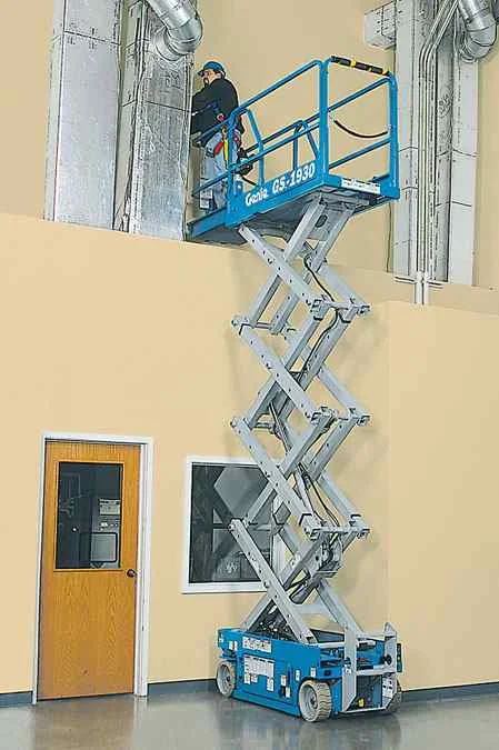 Ausgefahrene blaue Arbeitsbühne mit Person vor Rohren an heller Wand, daneben eine Tür.